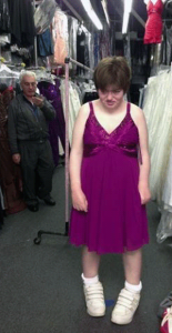 Siobhan in a purple old-lady dress with racks of clothes and a guy talking on the phone in the background.